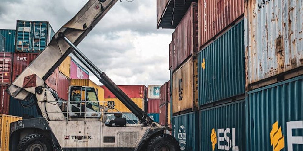 A crane loader stacking colorful shipping containers at a port with reflection in water.
